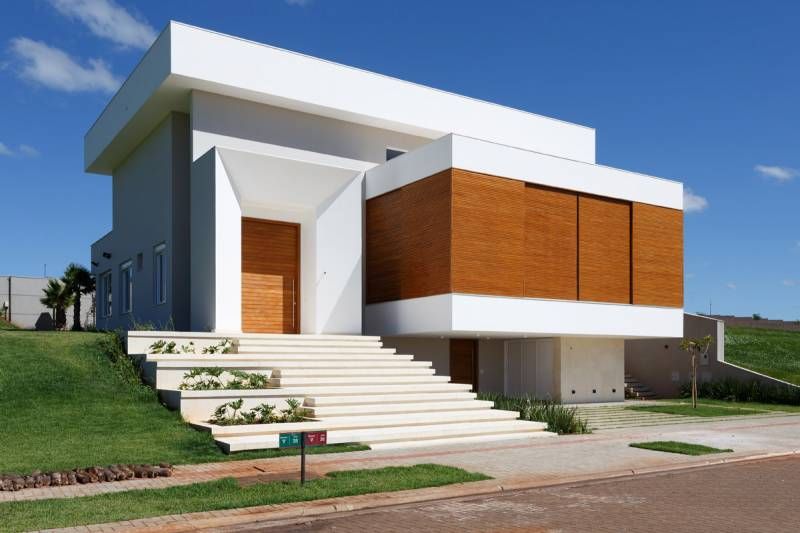 Casa moderna de dois andares com exterior branco e de madeira, escadas que levam à entrada, situada em um gramado verde sob um céu azul.