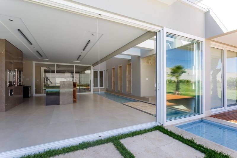 Casa moderna de conceito aberto com paredes de vidro, piscina e piso bege. No exterior, a luz do sol reflete nas janelas.