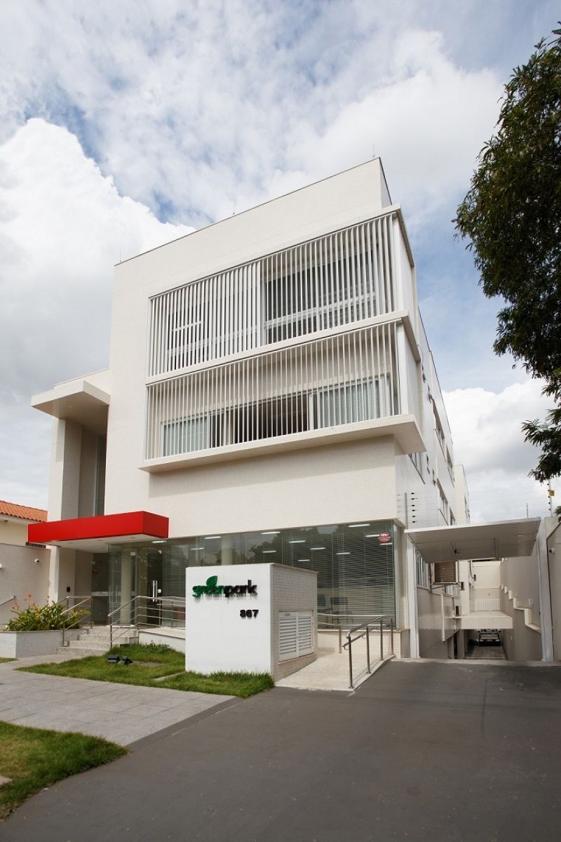 Prédio branco com detalhes em vermelho, placa verde e persianas verticais.