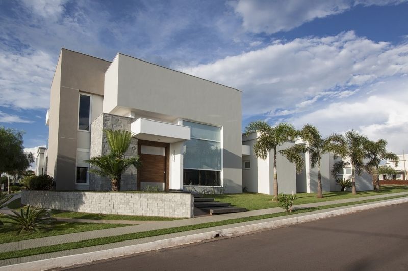 Casa moderna de dois andares com telhado plano e exterior cinza claro, palmeiras e céu azul.