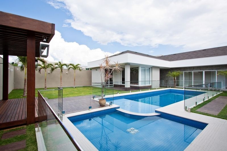 Quintal com uma piscina de azulejos azuis, gramado verde, um deck e uma casa branca moderna sob um céu nublado.