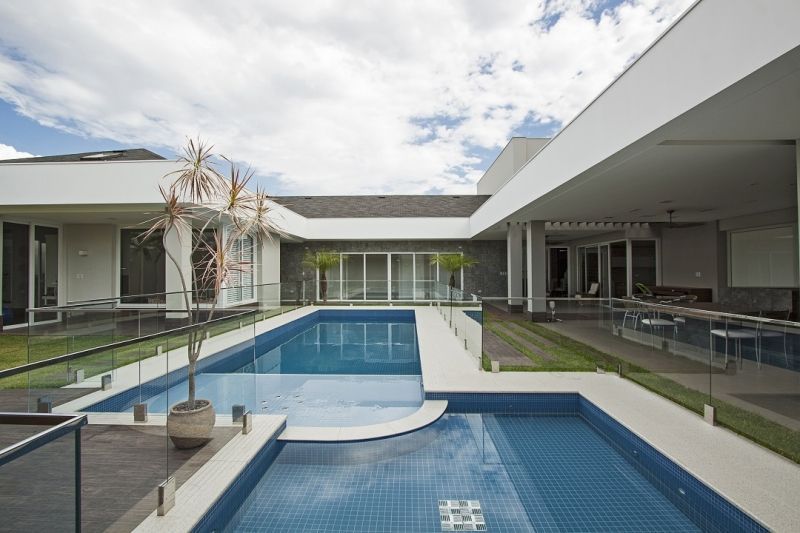 Casa moderna com piscina; grades de vidro, paredes brancas, água azul, dia ensolarado.
