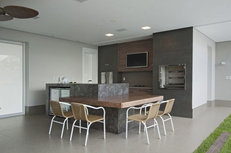 Cozinha ao ar livre com churrasqueira, mesa, cadeiras, TV e geladeira pequena, em um prédio moderno.