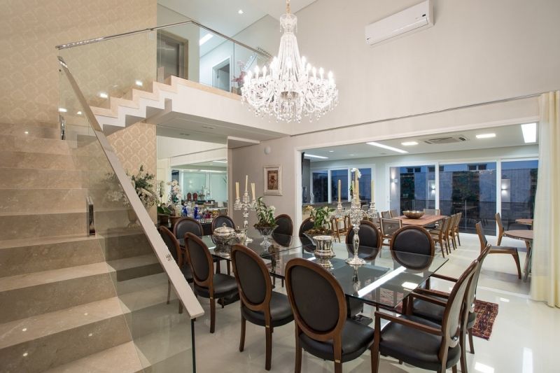 Elegante sala de jantar com mesa de vidro, cadeiras pretas e lustre de cristal. Escada para o andar superior.