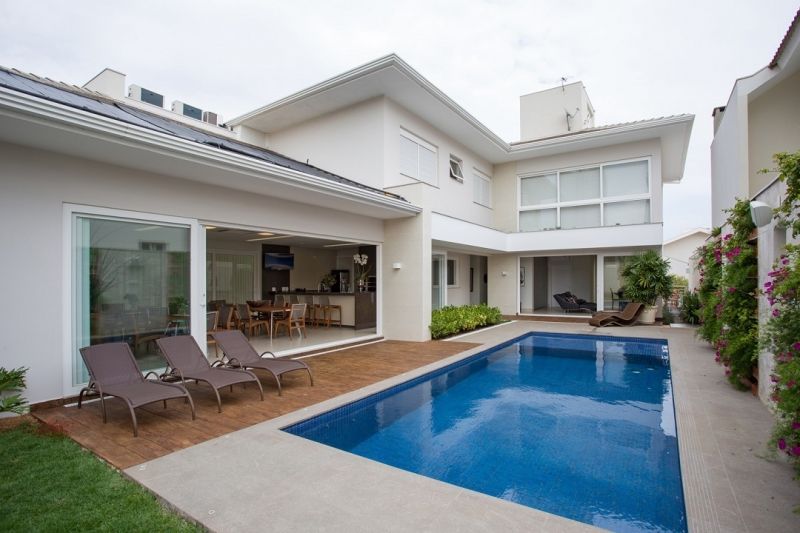 Casa de dois andares com piscina, deck e área externa com mesas. Exterior branco, água da piscina azul e gramado verde.