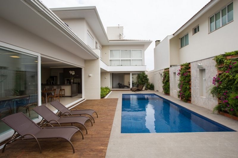 Quintal com piscina, espreguiçadeiras e casa branca de dois andares com deck de madeira e plantas trepadeiras.