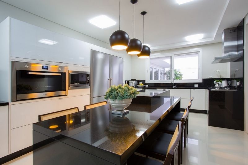 Cozinha moderna com ilha preta, armários brancos, eletrodomésticos de aço inoxidável e luminárias pendentes.