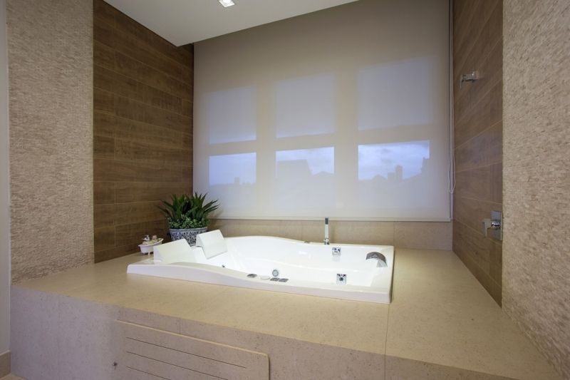 Banheiro moderno com banheira de hidromassagem branca embutida, paredes de cores claras e uma janela com persiana.