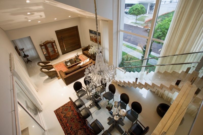 Vista de cima de uma sala de estar e jantar bem iluminada, com escada, lustre e janelas grandes.