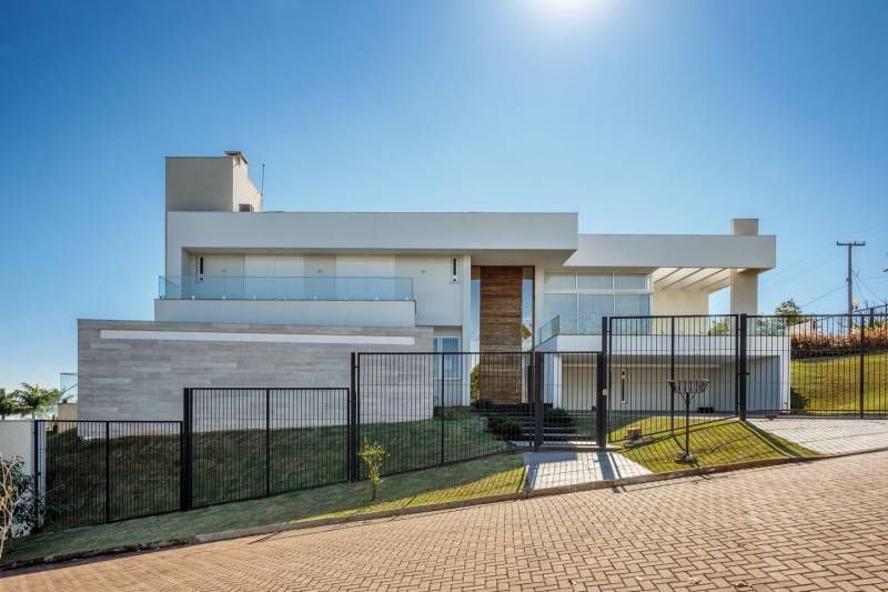 Casa branca moderna com grades de vidro, cerca escura e entrada de tijolos em um dia ensolarado.