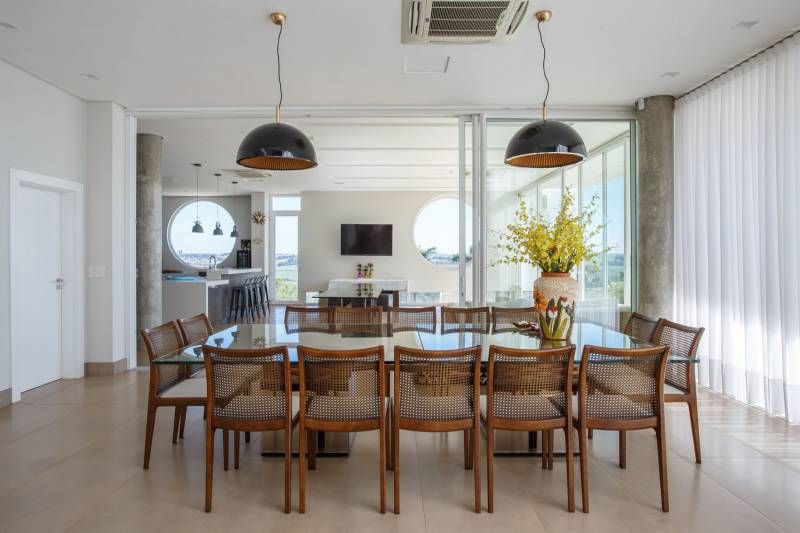 Sala de jantar com mesa grande, cadeiras e dois pendentes pretos. Vista da cozinha através das portas de vidro.