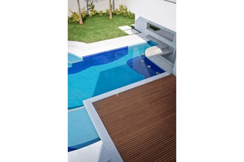 Piscina externa moderna com azulejos em tons variados de azul. Deck de madeira e gramado verde ao fundo.