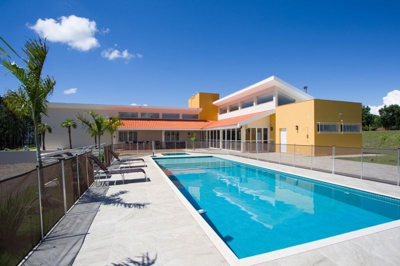 Uma piscina retangular em frente a um prédio amarelo com um teto laranja sob um céu azul.