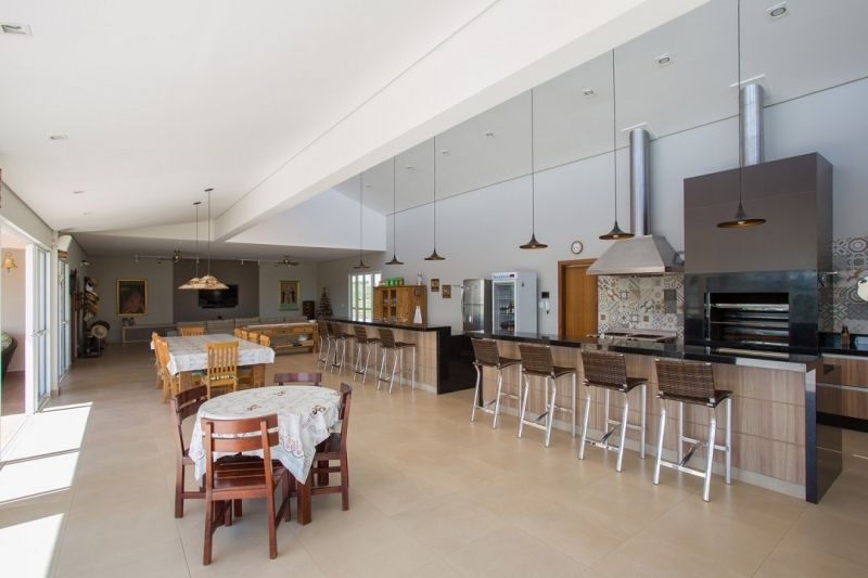 Espaço interno amplo e iluminado, com mesas de jantar, bancos de bar e churrasqueira. Cores neutras e muita luz.