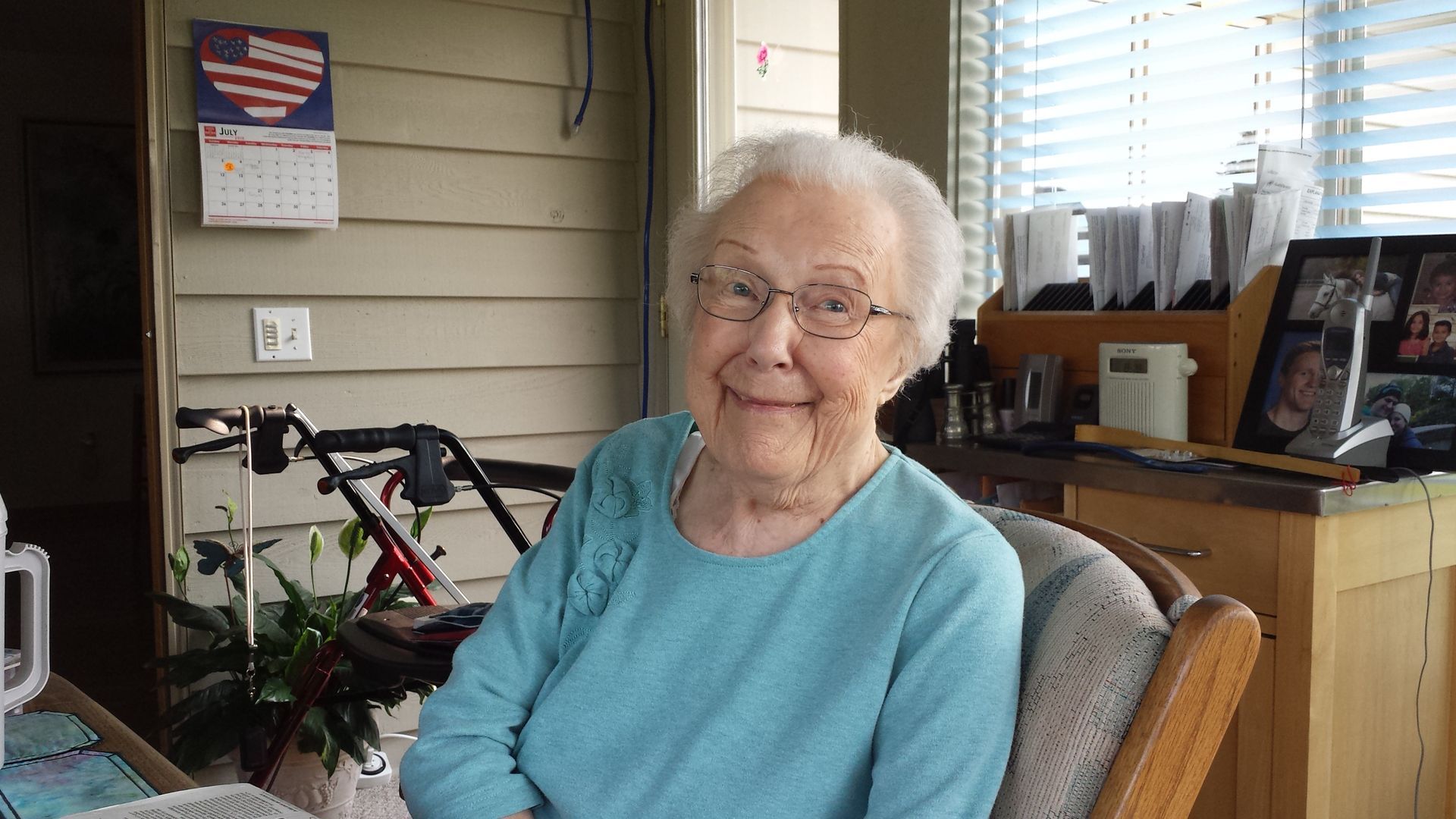 Elderly woman in a blue shirt and glasses smiles, sitting inside with a walker and desk visible.