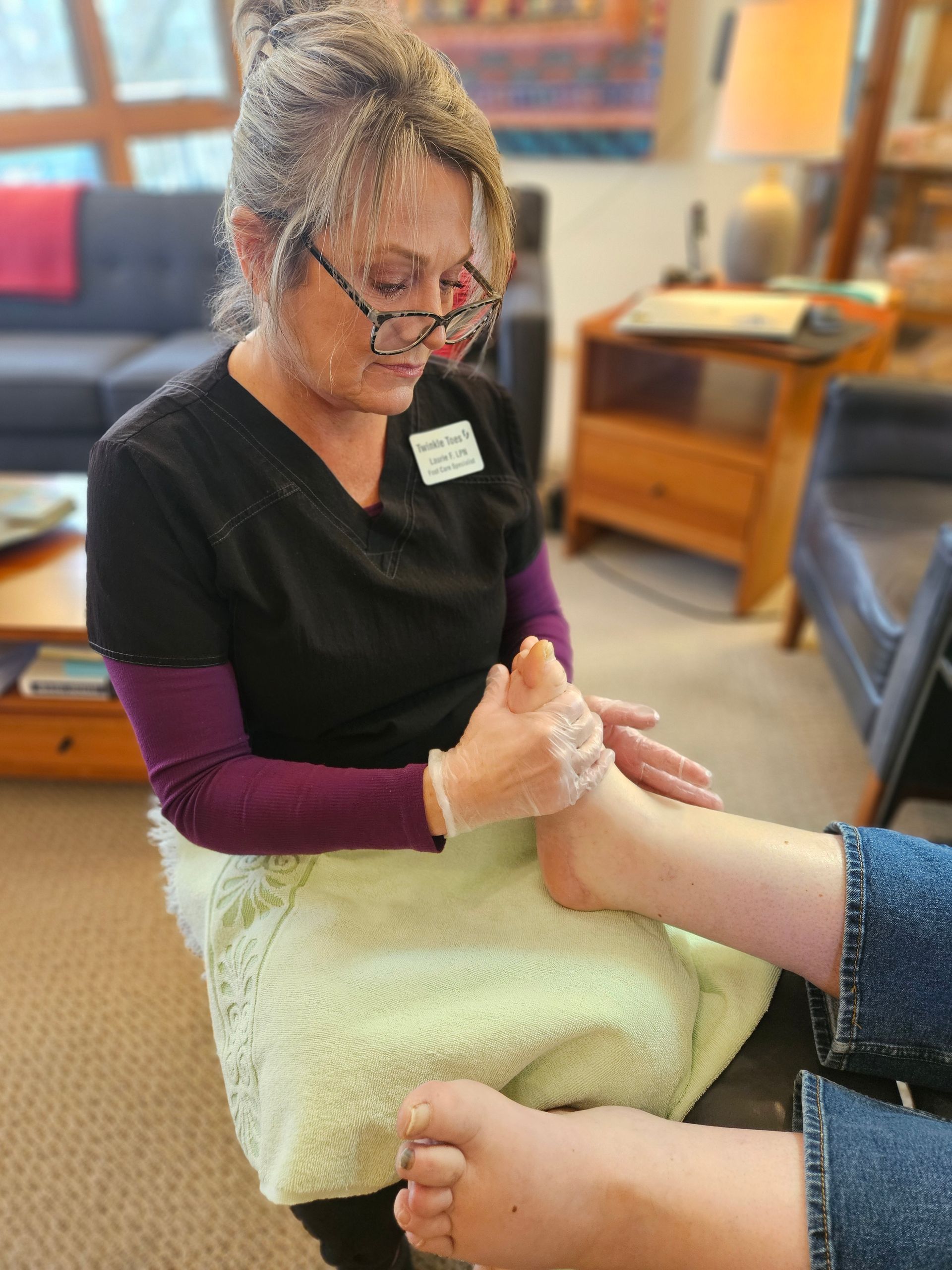 A caregiver wearing scrubs and gloves performs a foot massage for a person in a living room setting.