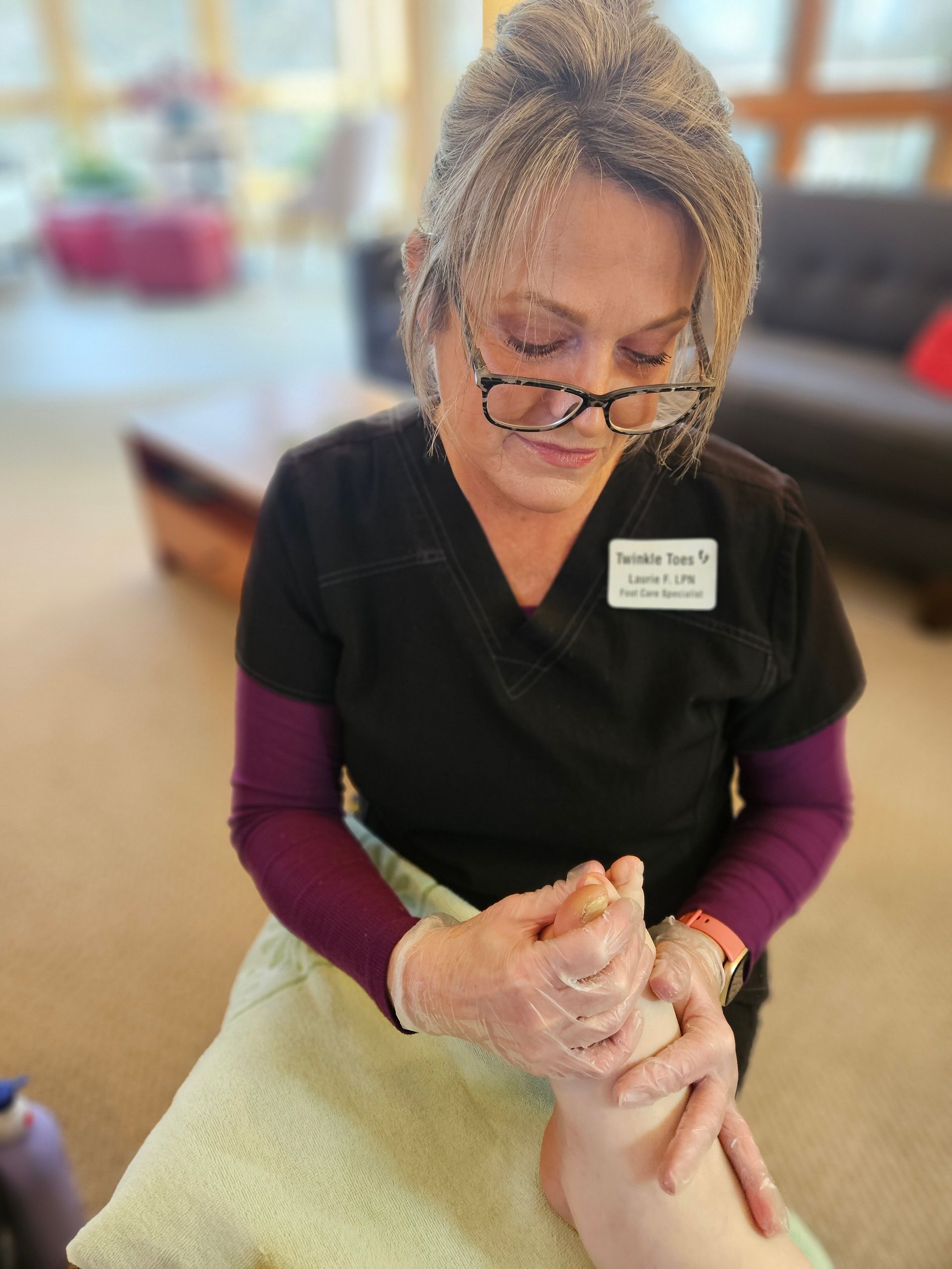 A professional wearing a name tag and gloves performs a foot massage in a room with a couch and blurred background.