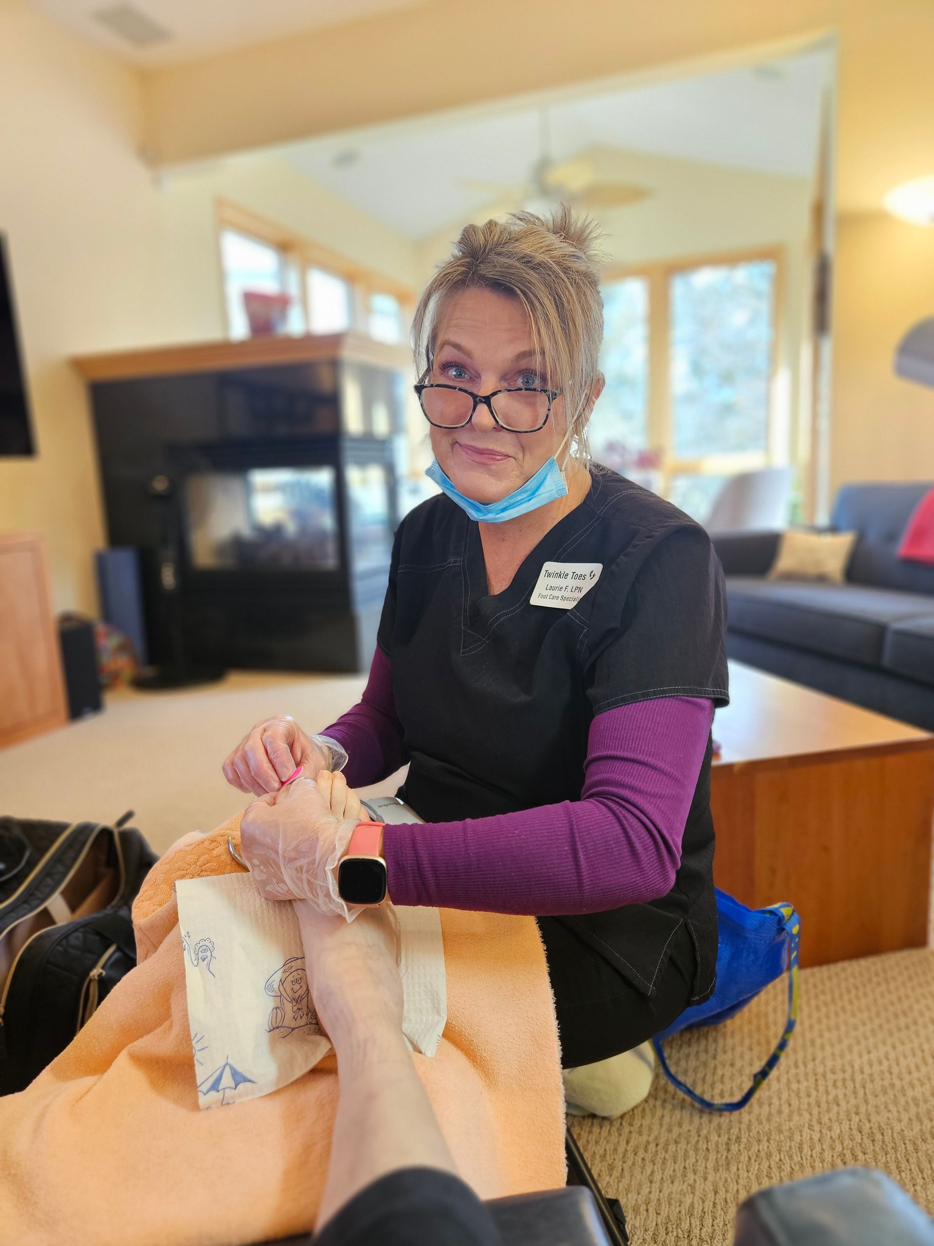Laurie, wearing scrubs, glasses, and a face mask, examines a person’s foot resting on an orange towel indoors.