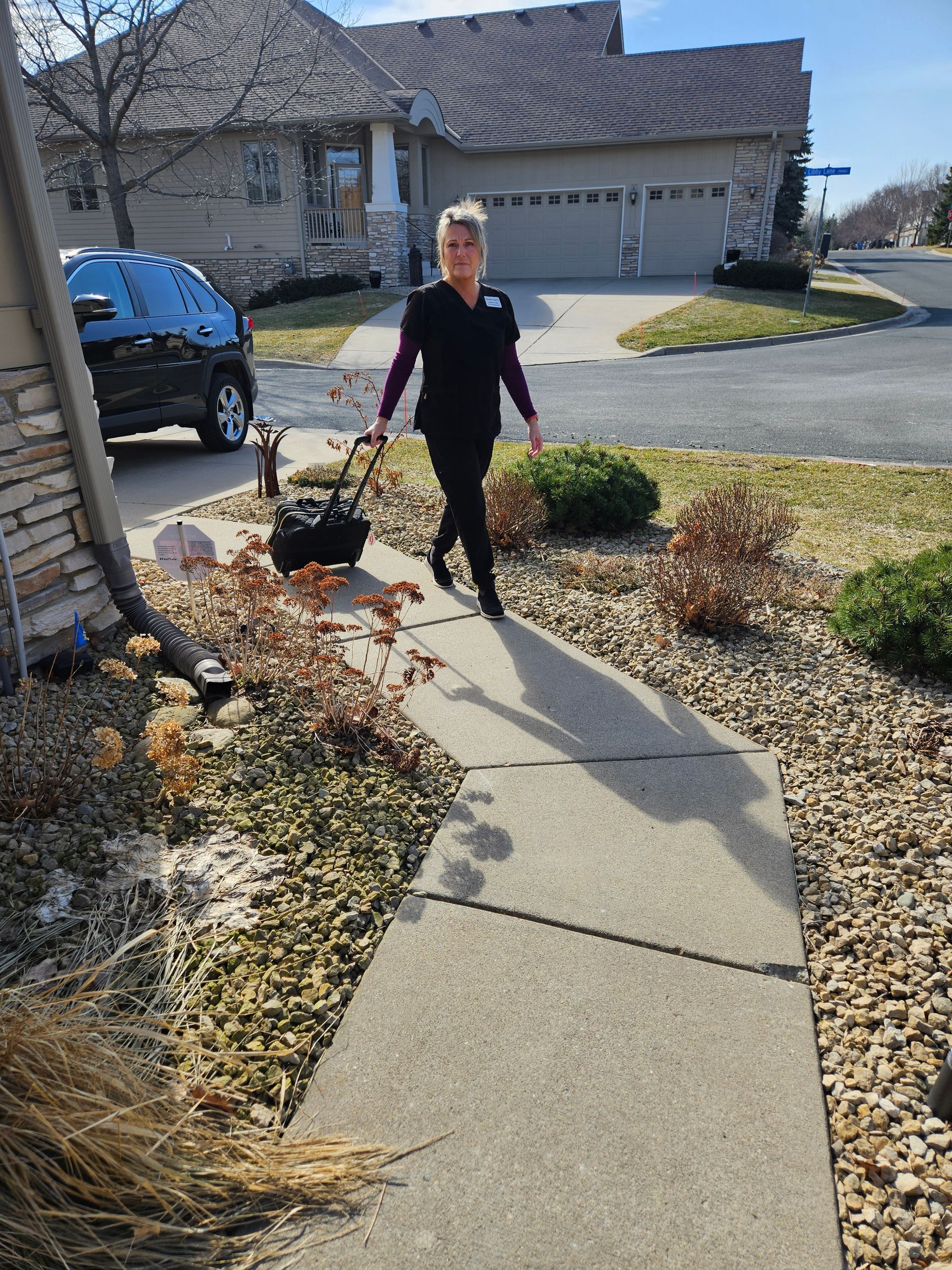 Laurie, Twinkle Toes, in dark scrubs, pulling a wheeled medical bag, walks up a paved path toward a suburban house.