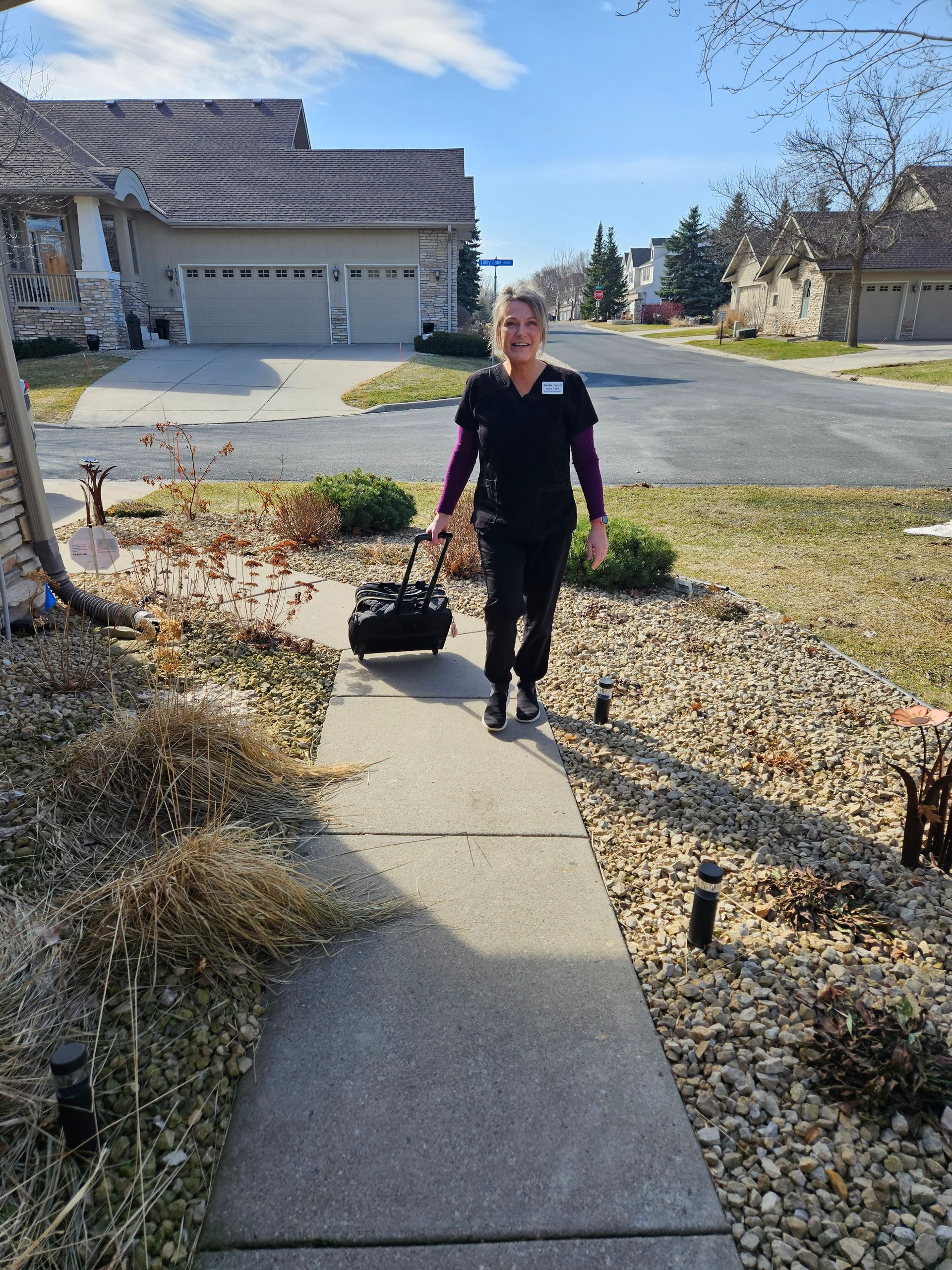 Laurie, Twinkle Toes, in black medical scrubs, walking up a residential sidewalk, pulling a small black equipment bag on wheels.