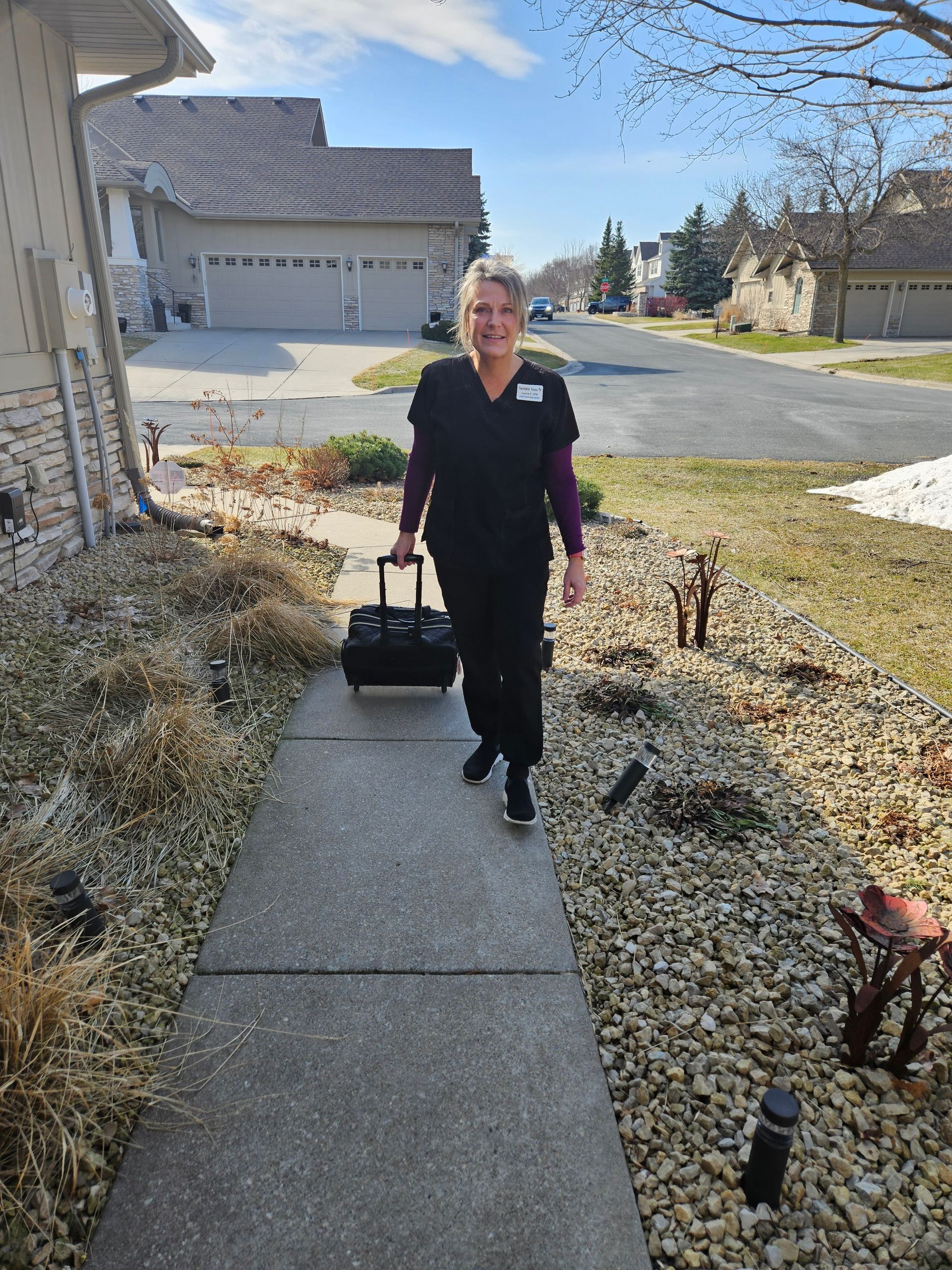 Laurie, Twinkle Toes, in black scrubs, walking down a suburban sidewalk, pulling a black rolling medical bag.