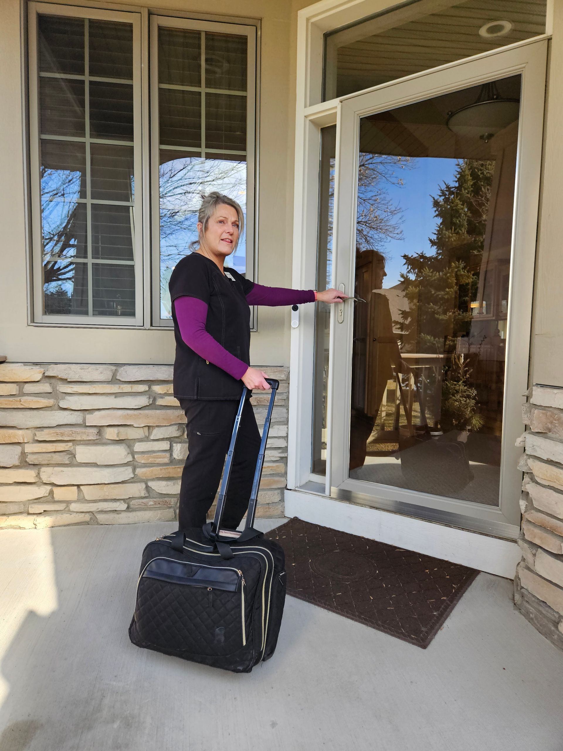 Laurie, Twinkle Toes, wearing dark medical scrubs, pulls a rolling equipment bag toward a residential front door.