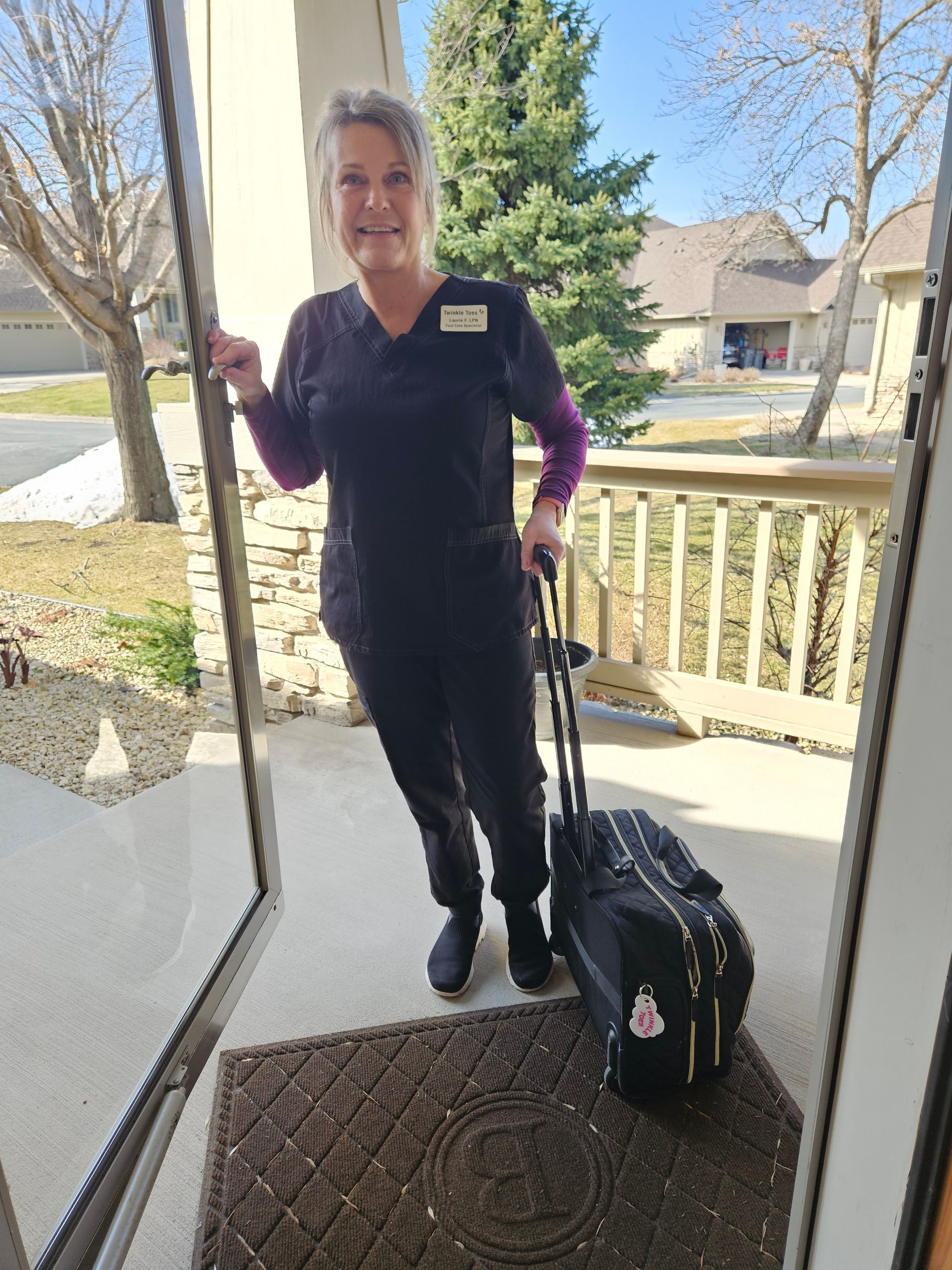 Laurie, Twinkle Toes, in black scrubs, stands in a doorway, holding a rolling medical bag, as seen from inside a house on a sunny day.