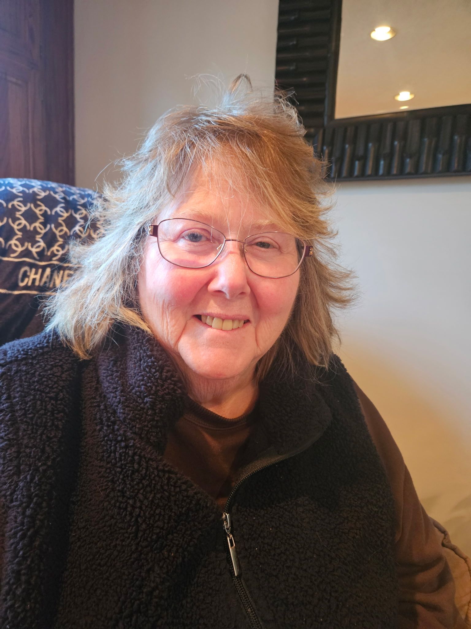 A smiling person with shoulder-length wavy hair and round glasses, wearing a dark brown zip-up fleece vest.