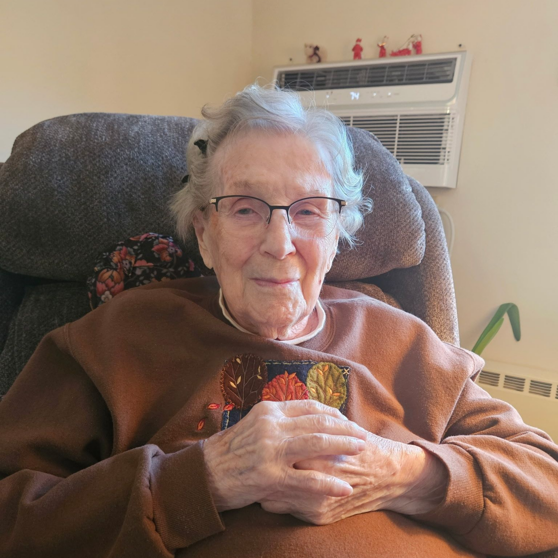 Elderly person in a brown sweater and glasses sits in a recliner, hands clasped.