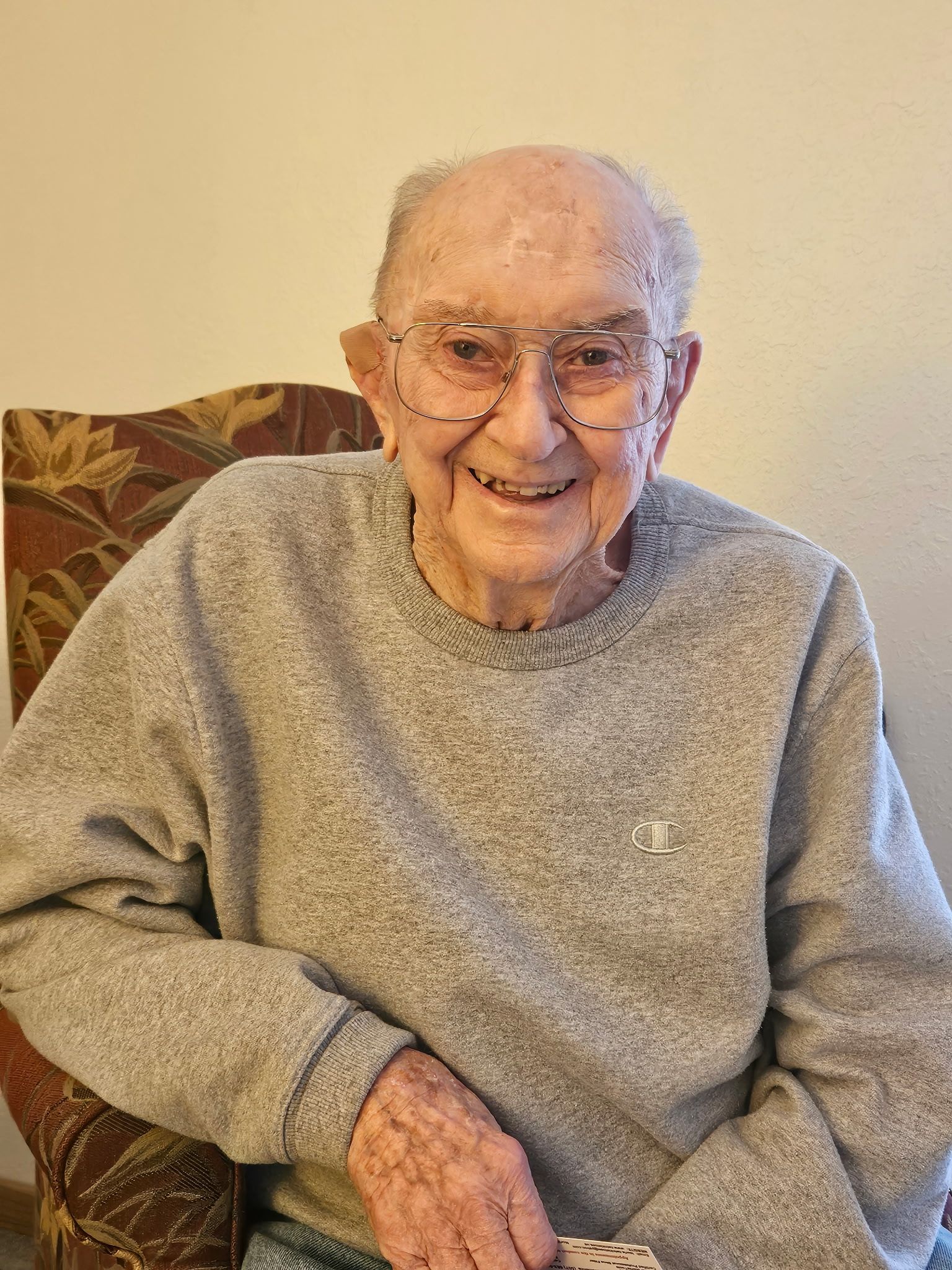 Smiling older person wearing glasses and gray sweater, sitting in a floral patterned chair.
