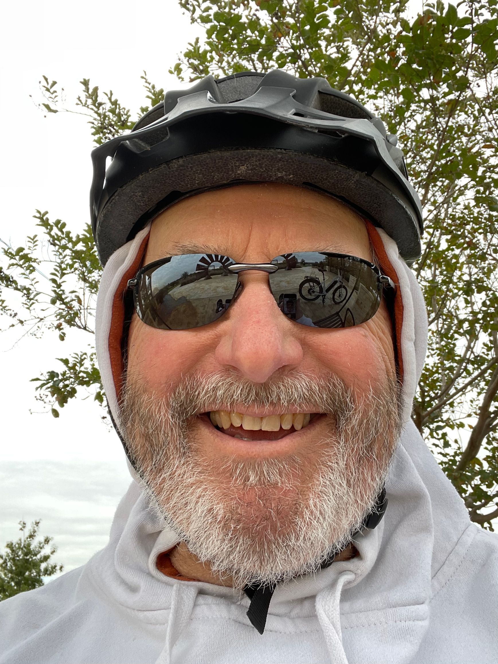 Smiling person in a cycling helmet, sunglasses, and a hooded sweatshirt with a frosted beard, outdoors against trees.