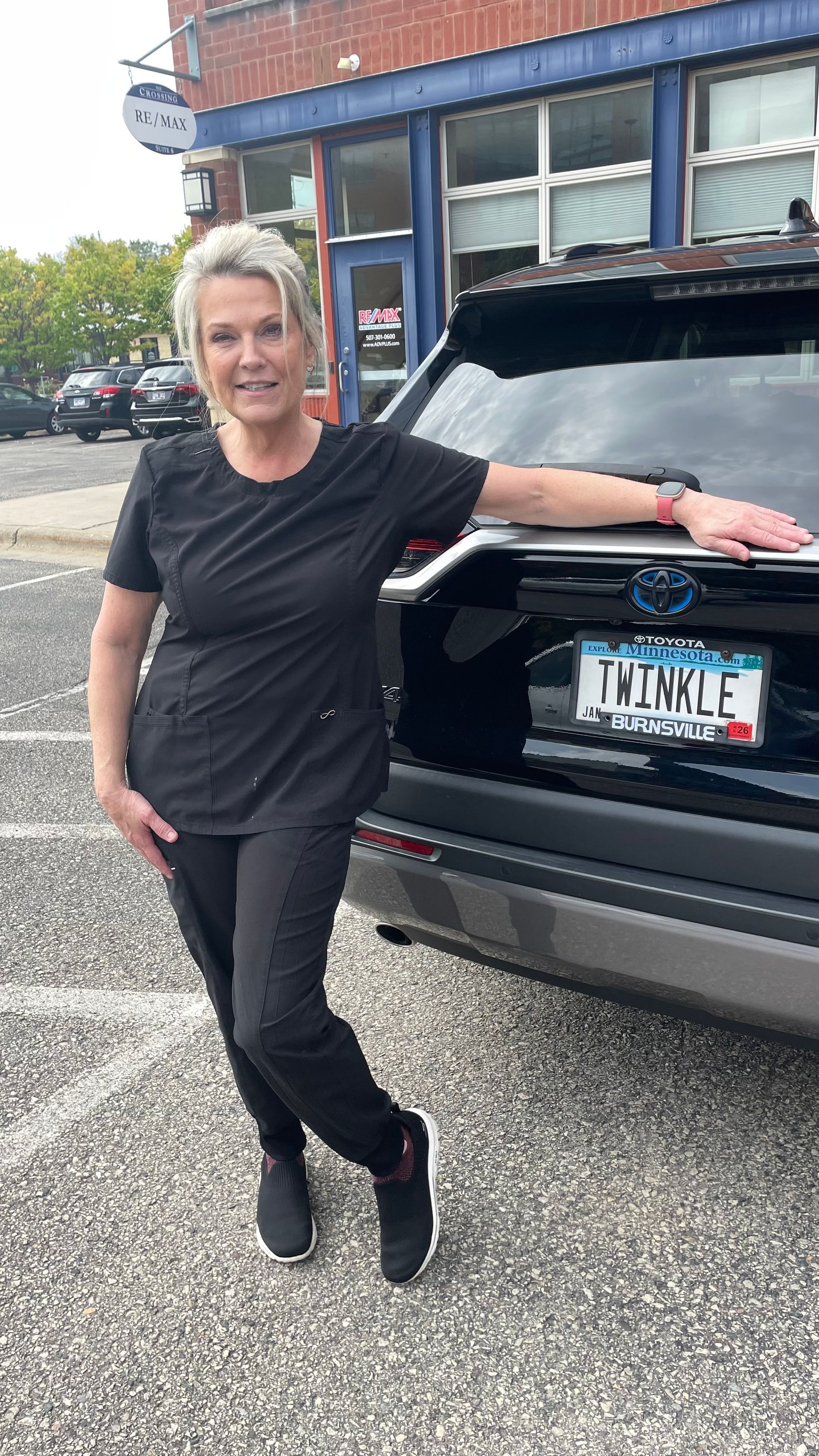 Woman in black scrubs is leaning on the back of a black SUV in front of a building.