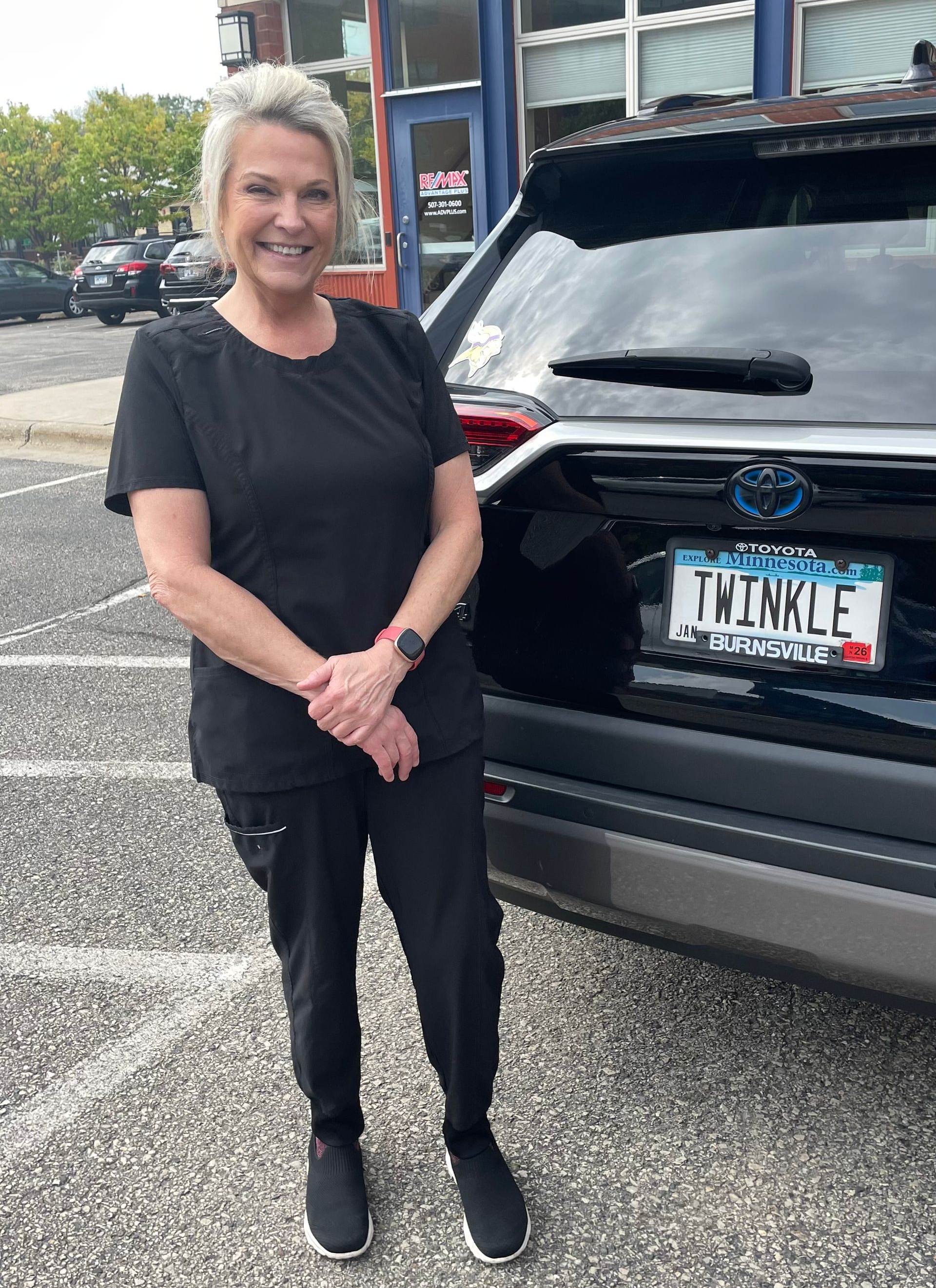 Woman in black scrubs, smiles, stands beside a black SUV with 