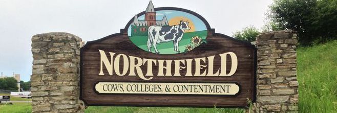 Sign for Northfield, featuring a cow, colleges, and contentment, set in stone pillars and surrounded by greenery.