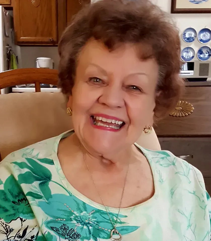 Elderly woman in a blue shirt and glasses smiles, sitting inside with a walker and desk visible.