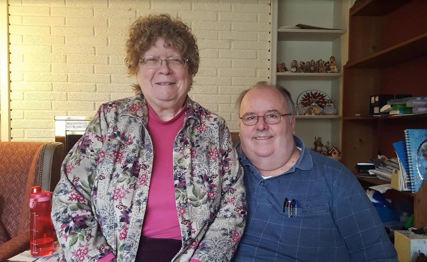 Two smiling people pose indoors, the woman wearing a floral jacket and the man wearing glasses and a blue shirt.