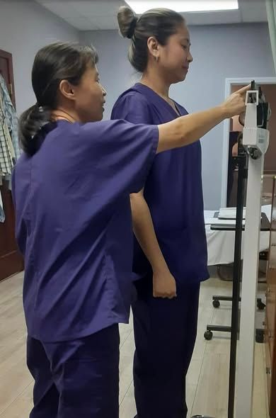 A person in purple scrubs adjusts a medical height measuring scale behind another person standing straight in the office.