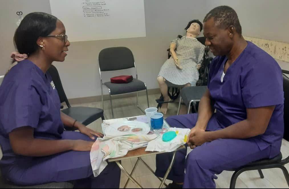 Two medical personnel in purple scrubs practice patient care techniques together at a table in a training classroom.