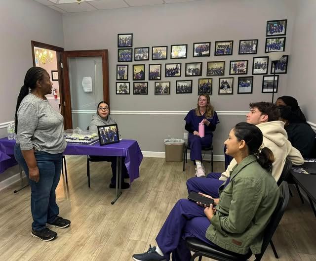 A group gathers in an office for a presentation, with a speaker standing near a table and attendees seated in chairs.