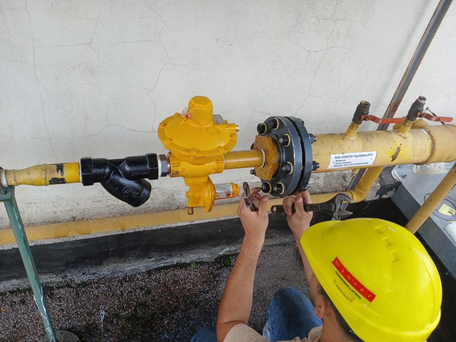 Un hombre con casco y mono inspecciona tuberías con una tableta en un entorno industrial.