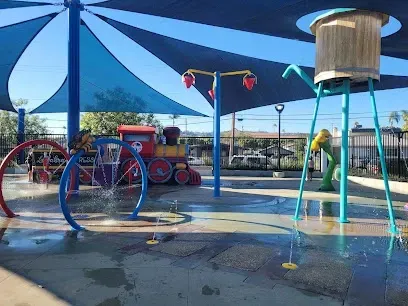 Tree Services West Covina -Children's splash pad with colorful water features, including giant bucket, train backdrop, and canopy. Sunny day, playful and lively atmosphere.