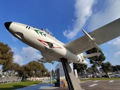 Tree Services West Covina -A white jet plane with red and green markings is mounted on display, angled upwards against a bright blue sky with fluffy clouds and surrounding trees.