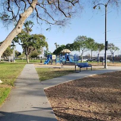 Tree Services West Covina-A sunny park with a winding path, nearby trees, and a blue playground set. Benches surround the area, creating a welcoming, serene atmosphere.