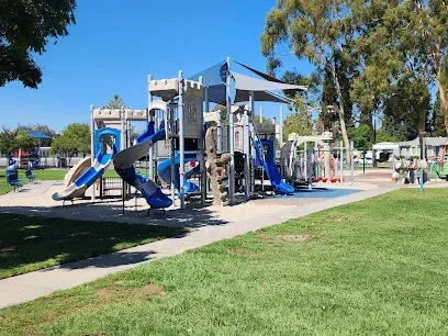 Tree Services West Covina Playground with blue slides and climbing structures under a shade canopy. Green grass and trees surround the area, creating a sunny, inviting atmosphere.