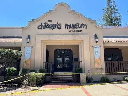 Tree Services West Covina -entrance of the Children's Museum at La Habra, featuring a beige facade with arched details, banners, and caution tape in front.