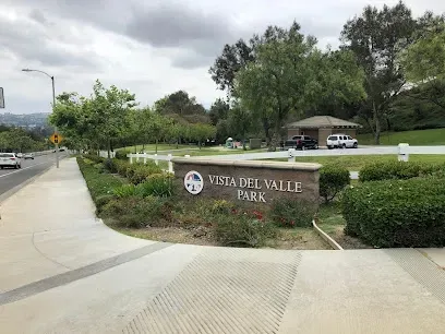 Tree Services West Covina-Vista Del Valle Park entrance with a stone sign, surrounded by greenery. A winding path leads into the park. Overcast sky and parked cars visible.