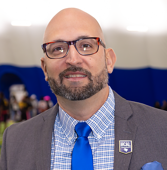 Man with crossed arms on a football field. He is wearing a blue polo shirt, khaki pants, and has a serious expression.