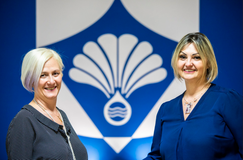 Due donne si stringono la mano davanti a un logo blu e bianco