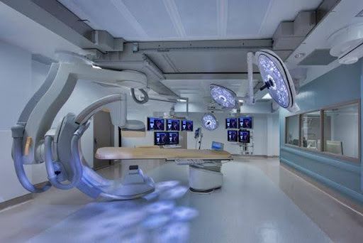 Uma sala de cirurgia moderna com equipamento médico especializado, luzes brilhantes no teto e vários monitores, sugerindo um procedimento em andamento.