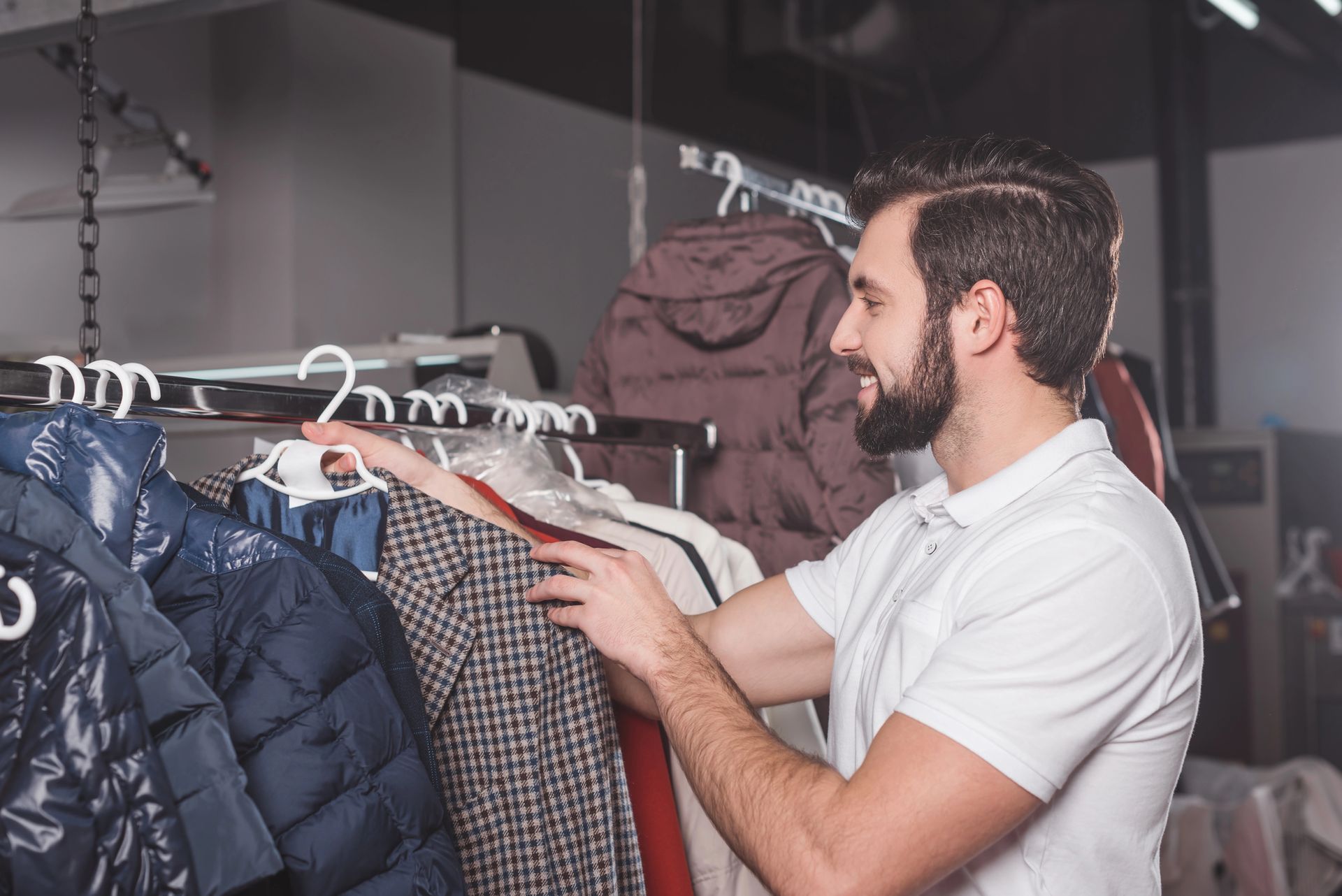 Una persona con barba que viste un polo blanco mira ropa colgada en un perchero en una tienda minorista.